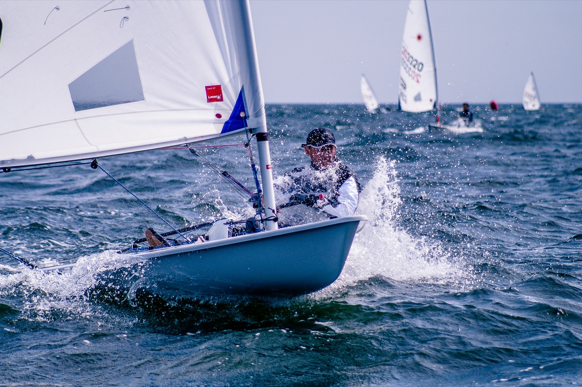 A Laser dinghy sailor driving through a wave upwind, spray flying